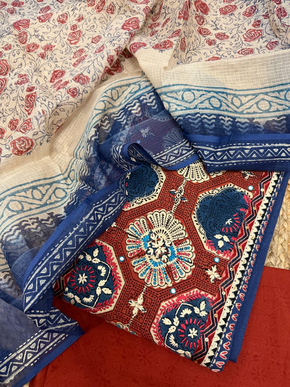 Navy blue and rust unstitched suit set – angled flat lay showing rust Ajrakh block print neckline panel with teal and white floral medallions, navy Kota dupatta border, and rust bottom fabric on rattan