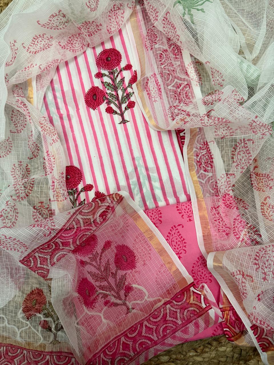 White and pink block print cotton 3-piece unstitched suit set – draped flat lay on rattan showing white stripe cotton kurta with red dahlia block print and red geometric arch border, pink paisley block print cotton bottom, and white Kota Doria dupatta with red floral bouquet and scallop arch border