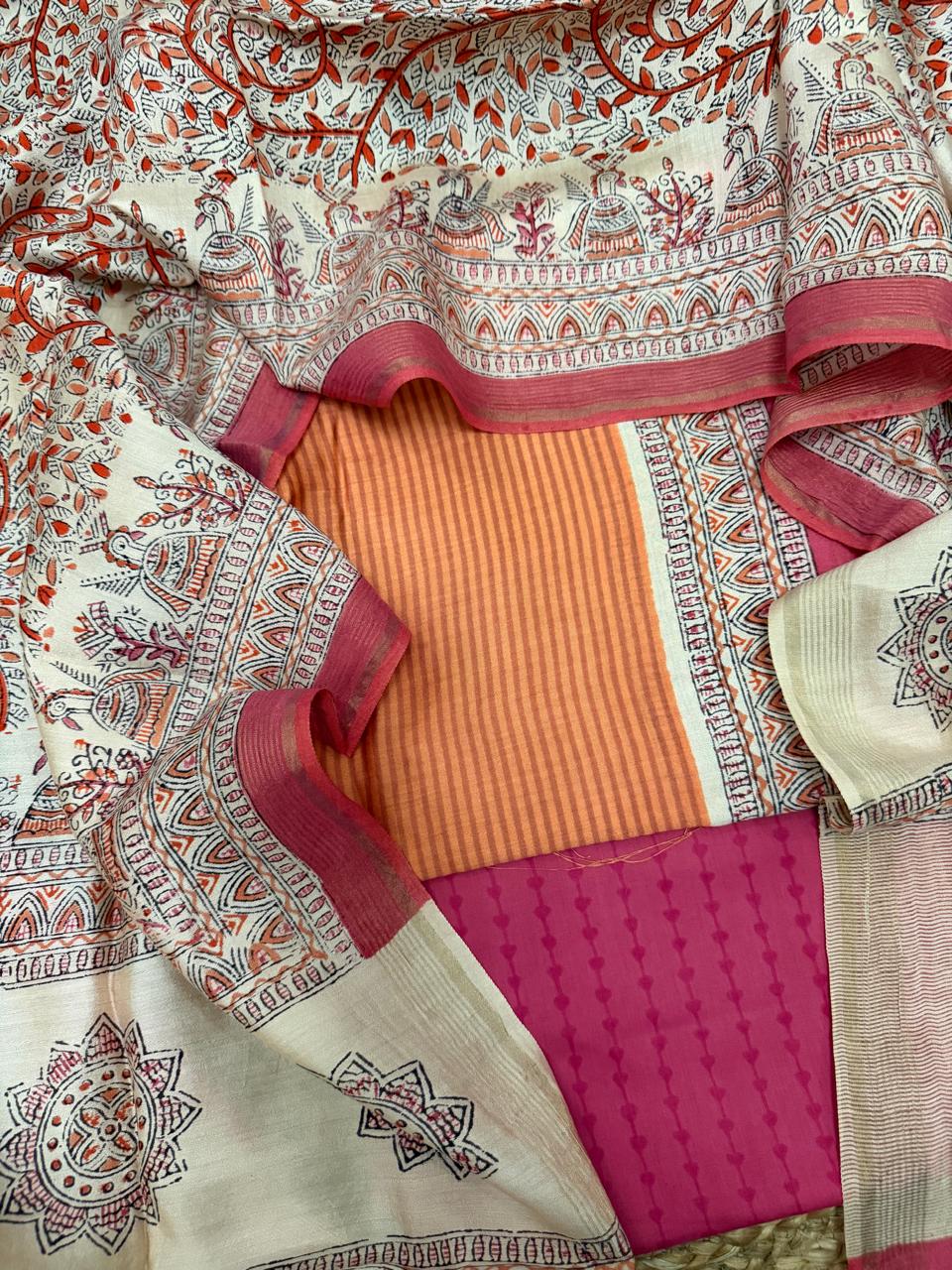 Orange and beige Maheshwari silk 3-piece unstitched suit set – overhead flat lay on rattan showing orange stripe kurta centred by cream Madhubani block print dupatta with red floral vine, temple arch border with peacock and diya motifs, and fuchsia diamond block printed bottom