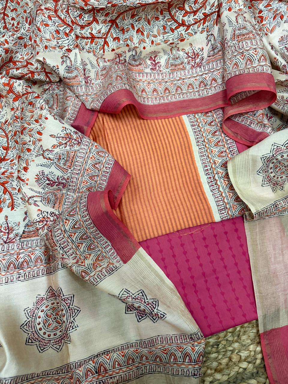 Orange and beige Maheshwari silk 3-piece unstitched suit set – angled flat lay on rattan showing orange stripe Maheshwari kurta framed by white Madhubani block print dupatta with red floral vine, temple arch border, peacock and diya motifs, and fuchsia diamond block printed bottom