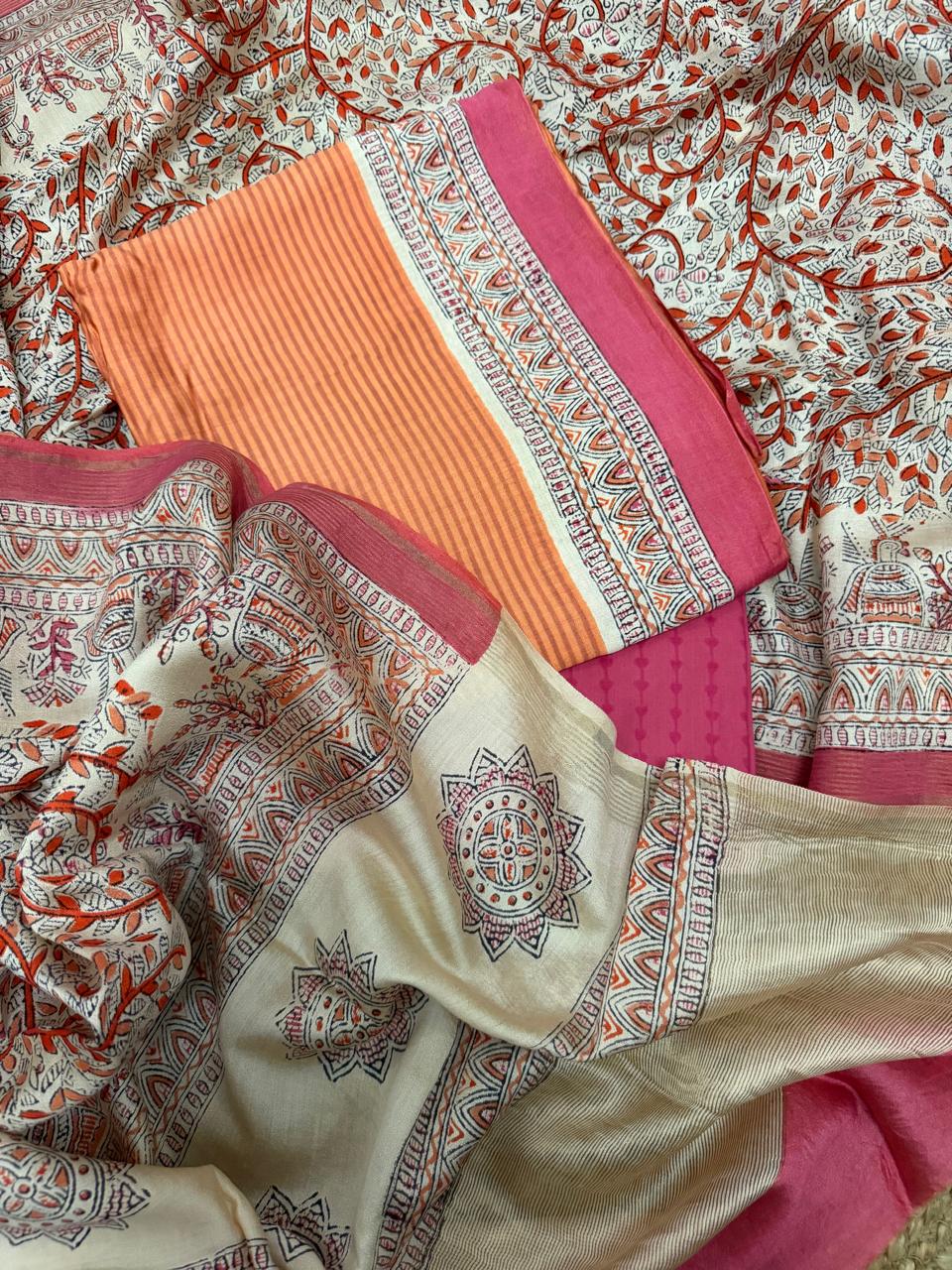 Orange and beige Maheshwari silk 3-piece unstitched suit set – draped flat lay showing orange stripe kurta, fuchsia diamond jacquard bottom, and cream Madhubani block print dupatta with red floral vine, black sun mandala butti, and multicolour temple arch peacock border