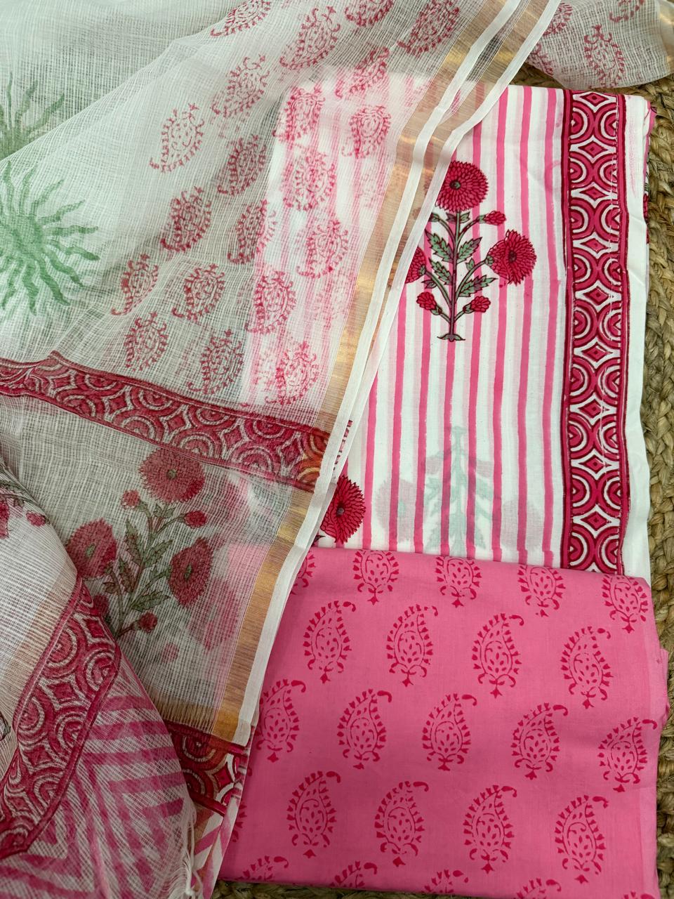 White and pink block print cotton 3-piece unstitched suit set – angled flat lay on rattan showing white Kota Doria dupatta with pink paisley butti and red dahlia floral bouquet alongside white stripe kurta with red dahlia print, red geometric arch border, and pink paisley block print cotton bottom