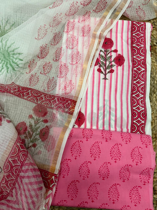 White and pink block print cotton 3-piece unstitched suit set – angled flat lay on rattan showing white Kota Doria dupatta with pink paisley butti and red dahlia floral bouquet alongside white stripe kurta with red dahlia print, red geometric arch border, and pink paisley block print cotton bottom