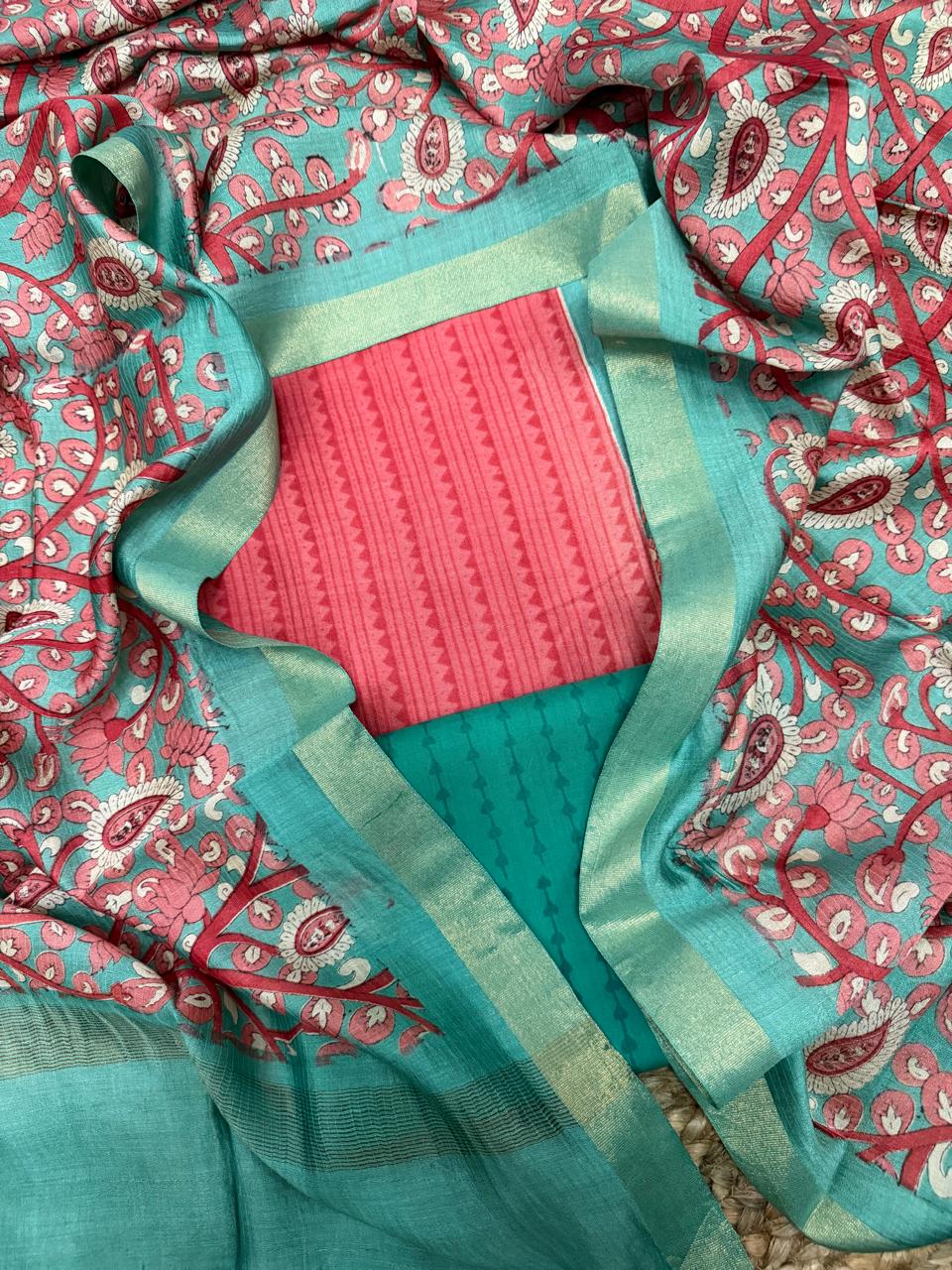 Teal and coral Maheshwari silk 3-piece unstitched suit set – draped flat lay on rattan showing coral stripe zigzag kurta framed by teal Kalamkari silk dupatta with pink lotus, paisley, and floral vine print, gold zari border, and teal dot-stripe cotton bottom