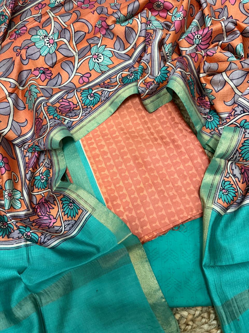 Orange and teal Maheshwari silk 3-piece unstitched suit set – overhead flat lay showing orange Kalamkari floral vine dupatta with teal zari border, orange geometric woven kurta fabric, and teal plain Maheshwari bottom